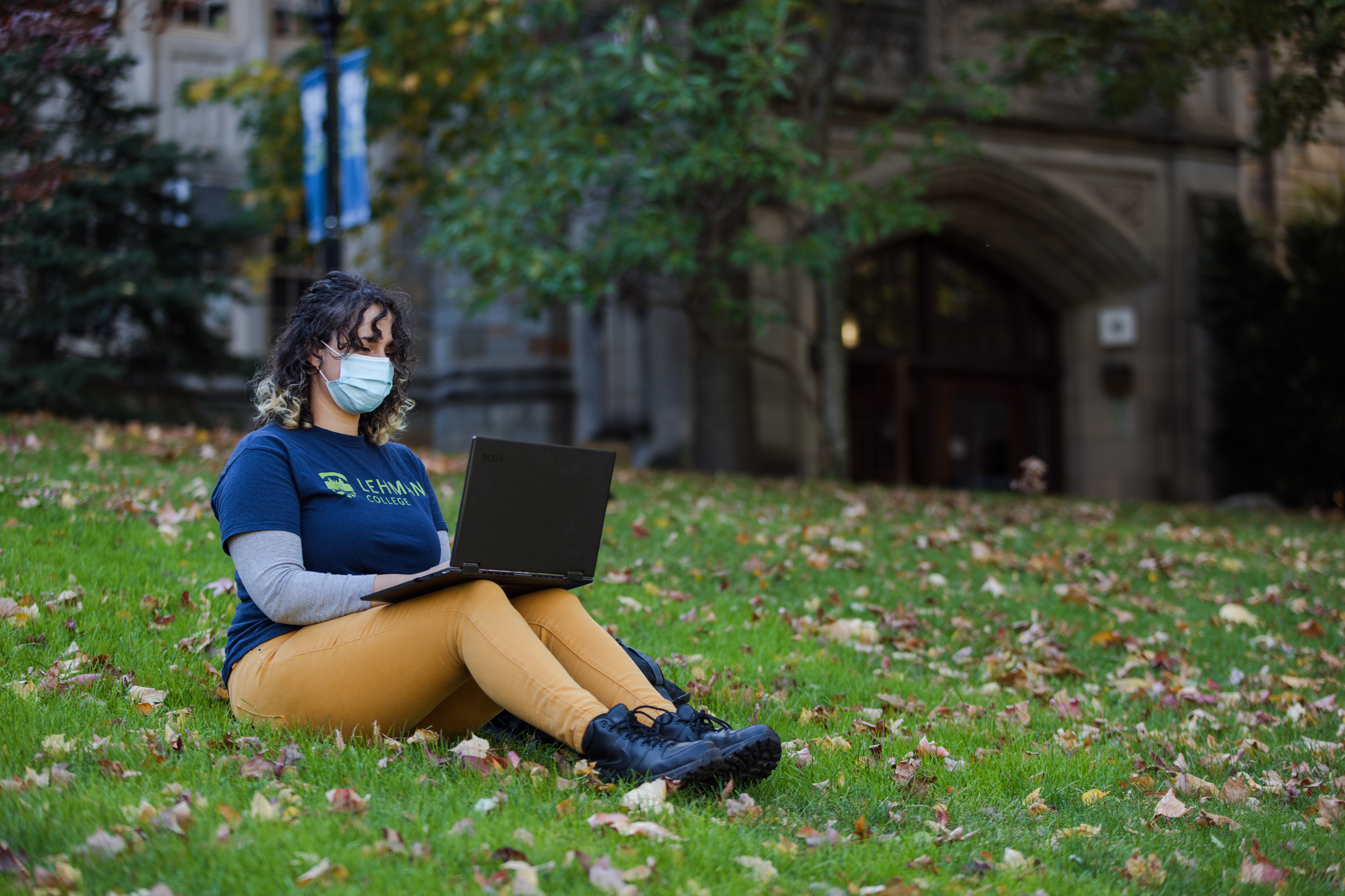 A young woman with a Lehman College T-shirt, masked and on campus, using a laptop