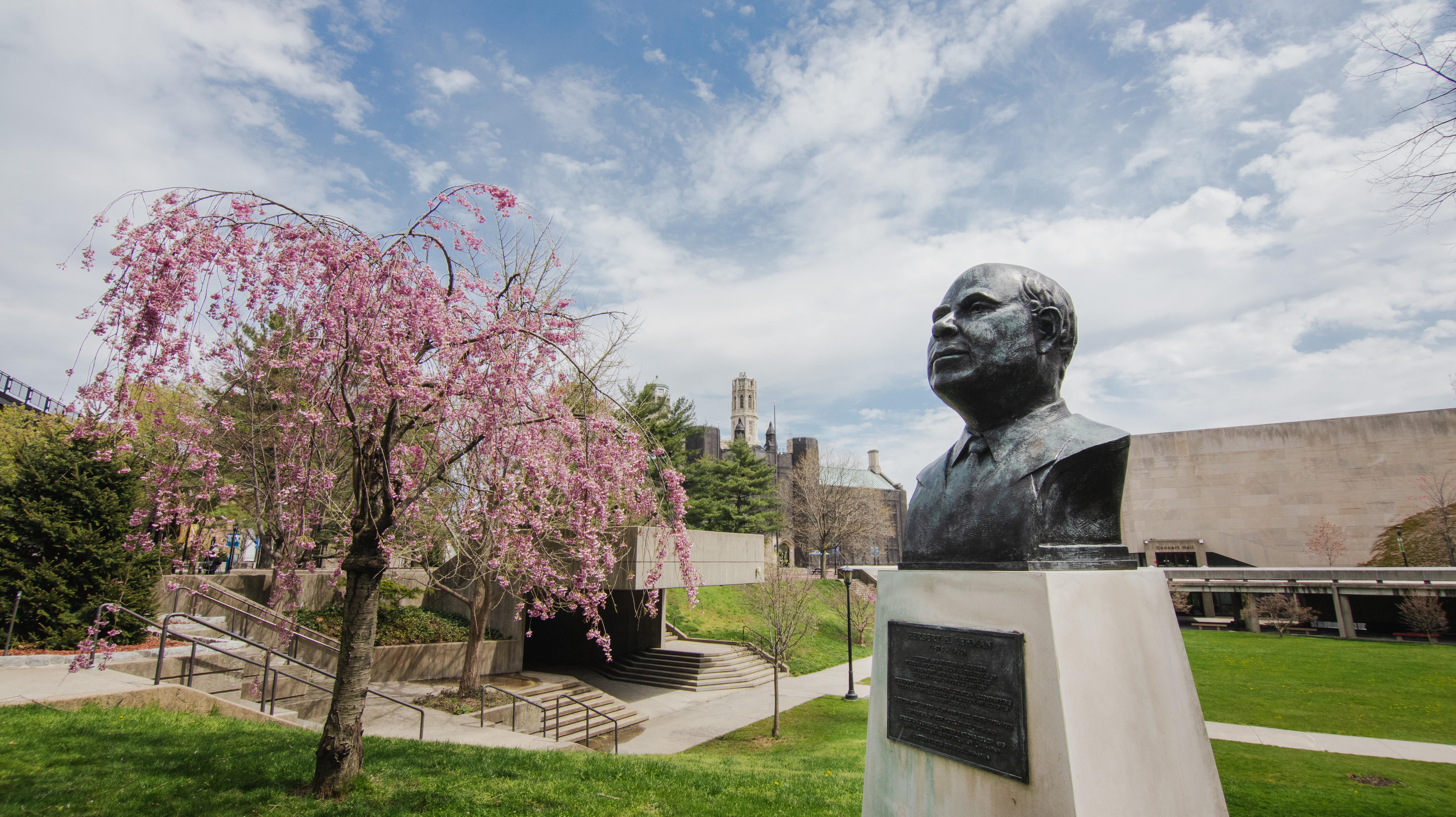 A view of the Lehman campus with a bust of Herbert Lehman in the right foreground with a blooming tree in the left background.