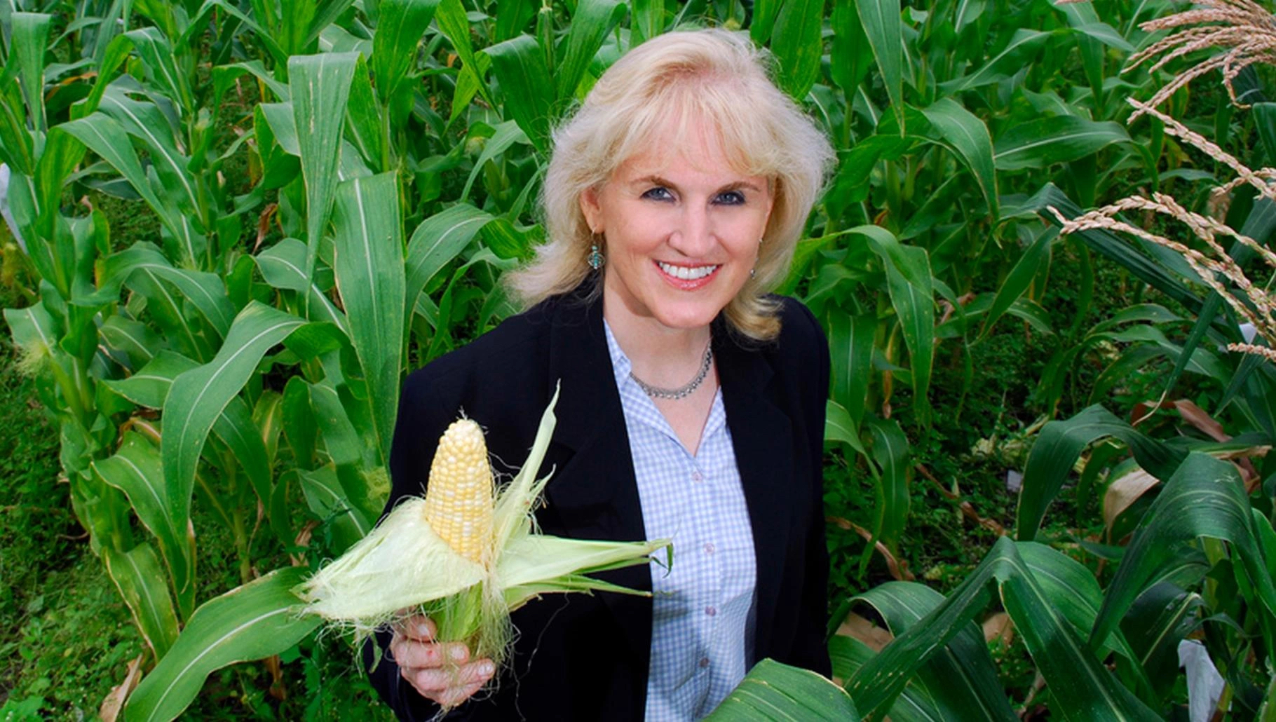 A blonde woman standing in a cornfield faces the camera, holding an ear of corn.