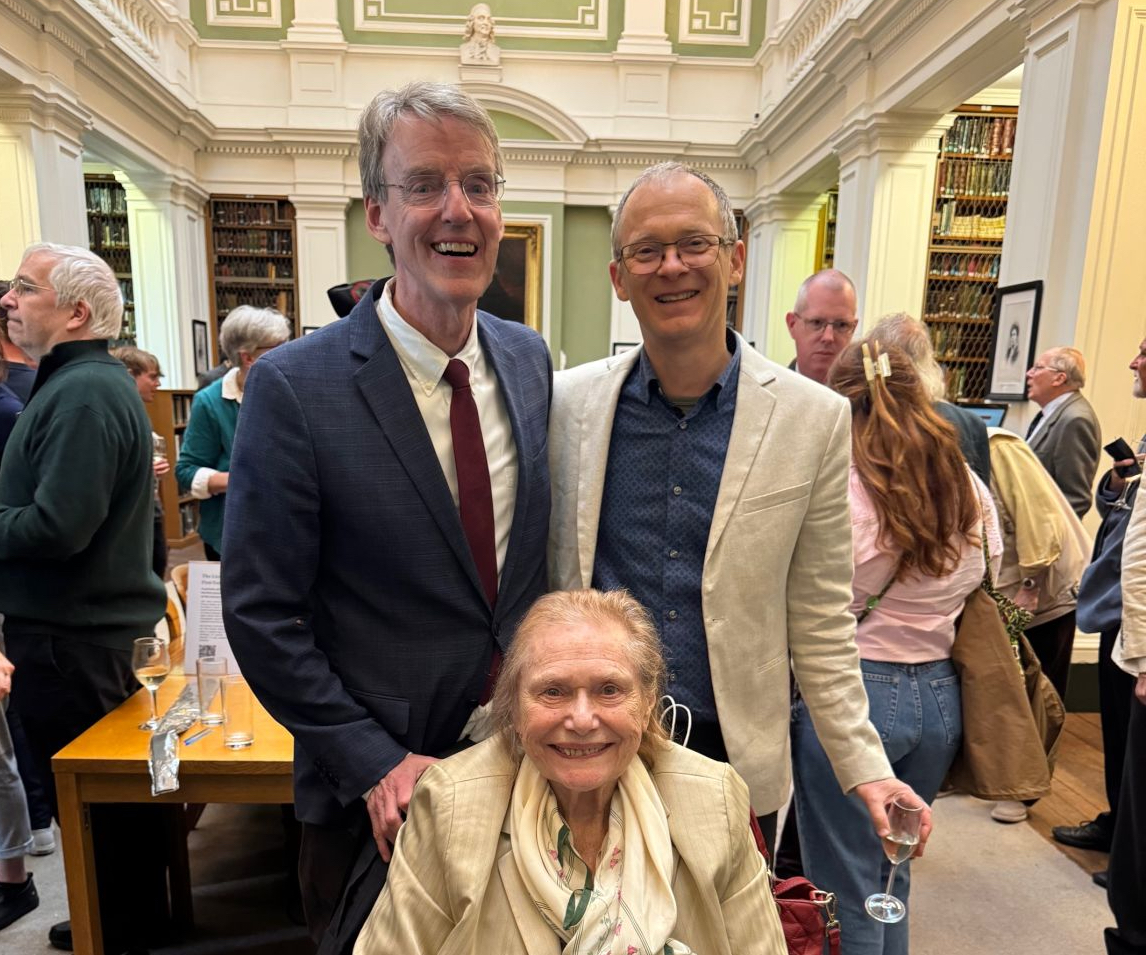 Snapshot of two men posing with an elderly woman in a wheelchair in a room of people