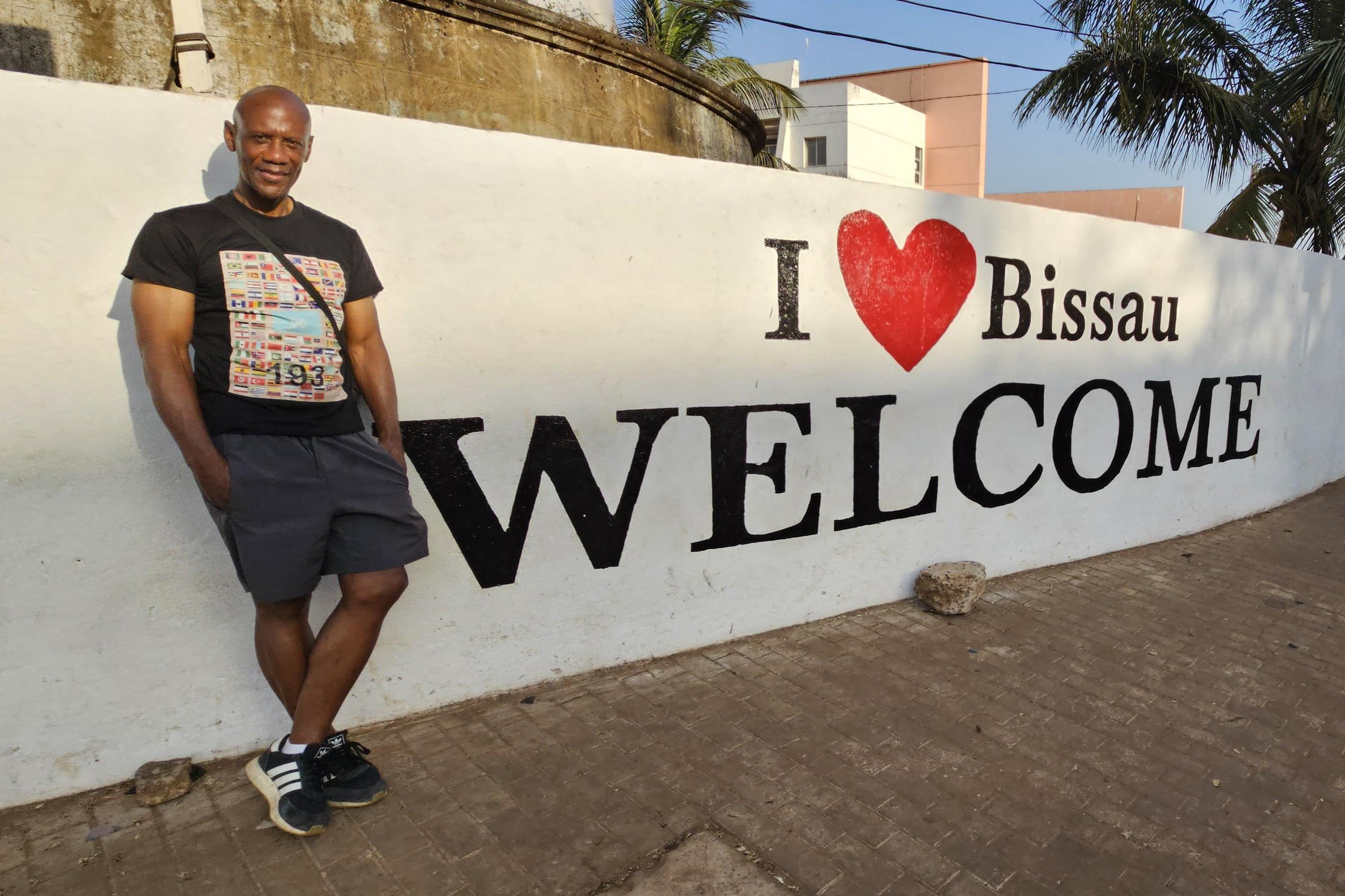 Bryan Warde stands next to a welcome sign painted on a wall in Guinea-Bissau.