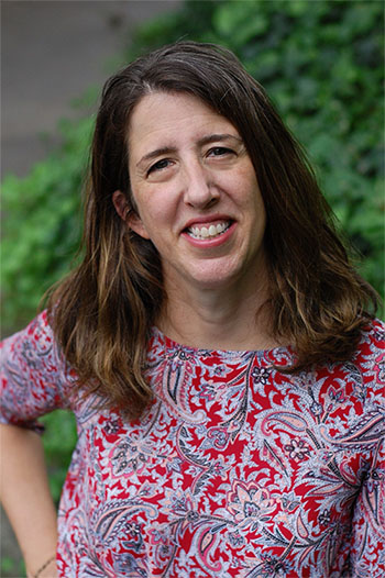 Woman with long brown hair wearing a red and white paisley-patterned top, standing outdoors in front of greenery and smiling at the camera.