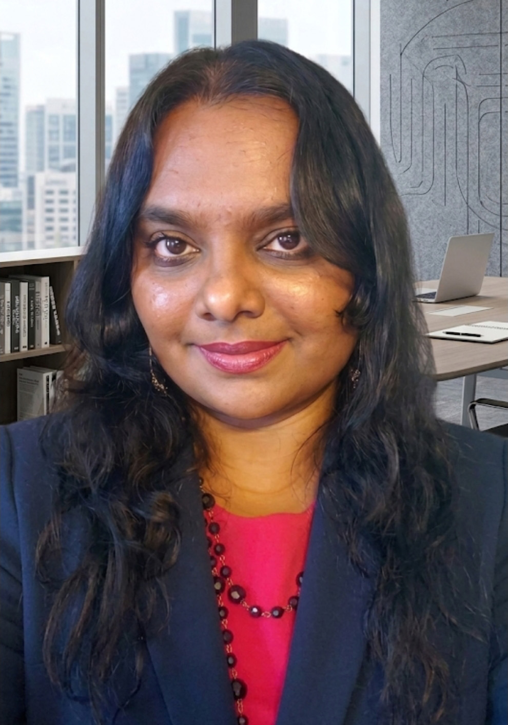 Professional headshot of a smiling person with long dark hair, wearing a dark blazer over a red top and a beaded necklace, with an office background featuring large windows, bookshelves, and a desk.
