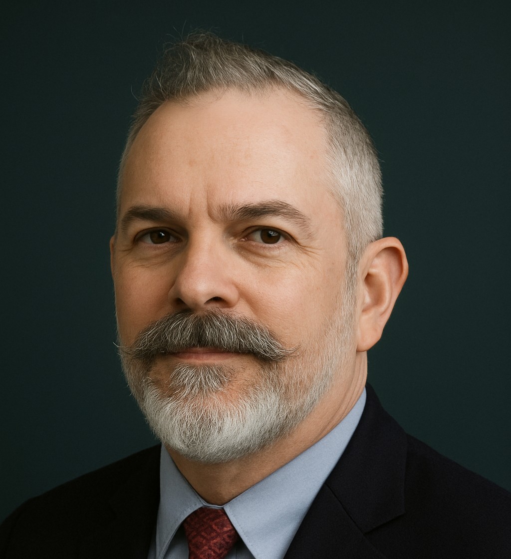 Professional headshot of a person with short gray hair and a gray beard and mustache, wearing a dark suit, light blue shirt, and burgundy tie, against a dark background.